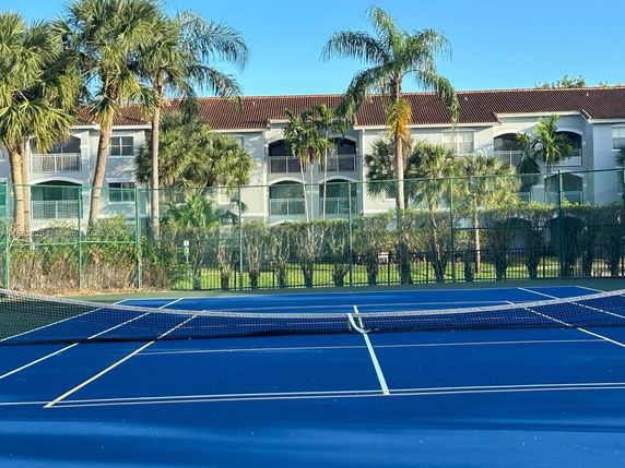 Tennis court with net and surrounding green fence, located near a building with balconies and palm trees.
