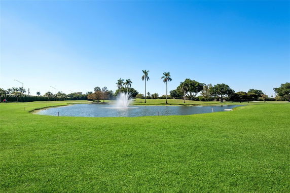 View of a grassy landscape with a pond and fountain surrounded by trees.