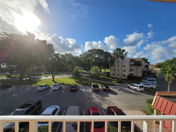 View of a parking area with multiple cars parked in rows, seen from a balcony. Residential buildings and trees in the background under a blue sky with clouds.