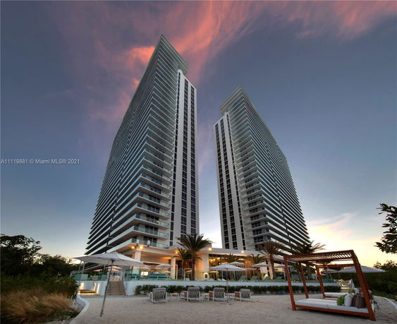 Front view of two tall residential buildings with multiple balconies and a surrounding recreational area.
