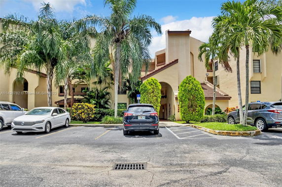 Outdoor parking area with cars in front of a multi-story building with arches and palm trees.