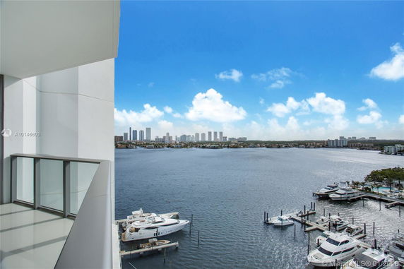 Panoramic view from a building balcony overlooking a body of water with yachts in a marina and a distant skyline of tall buildings.