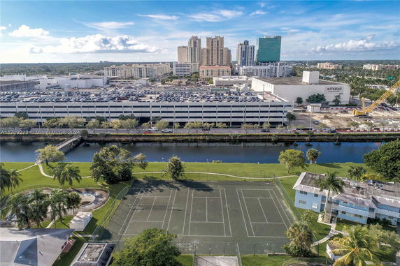 Aerial view of tennis courts adjacent to a canal, with a large parking structure and city buildings in the background.