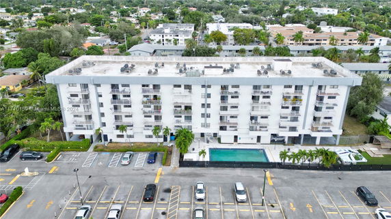 Aerial view of a multi-story residential building with parking area and swimming pool in front, surrounded by greenery and other buildings.
