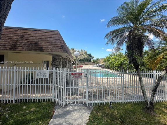 View of a swimming pool area enclosed by a white fence, adjacent to a small building with a stone facade and brown roof.