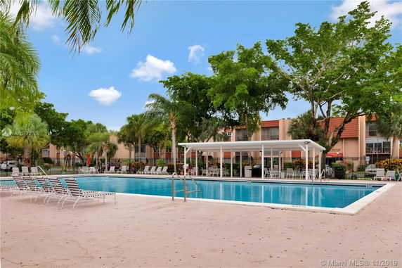 Outdoor swimming pool with lounge chairs and a shaded area, surrounded by trees and buildings.