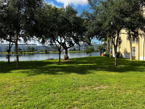 Outdoor view of a grassy area with trees next to a building, overlooking a lake with buildings across the water.