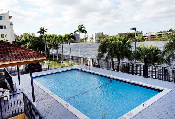 Outdoor swimming pool area adjacent to a building with a view of the water and palm trees.