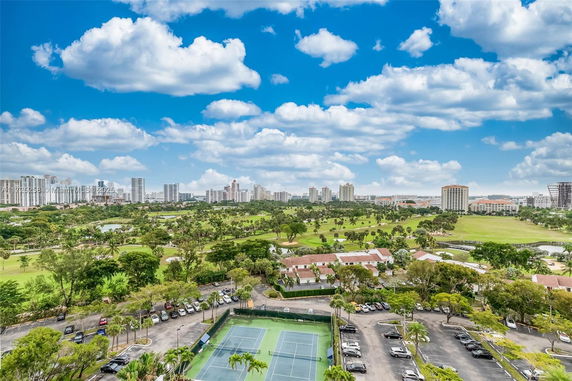 Wide view of a cityscape with tennis courts, parking area, and surrounding greenery.