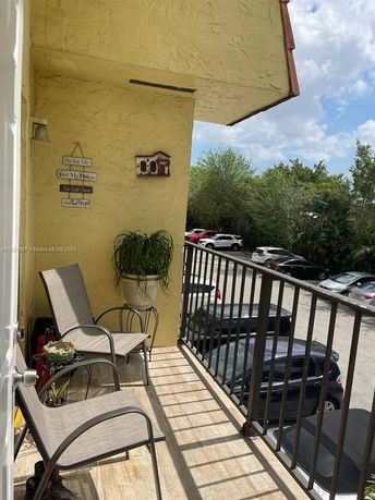 Outdoor view from a small balcony with chairs, potted plant, and railing overlooking parked cars and trees.