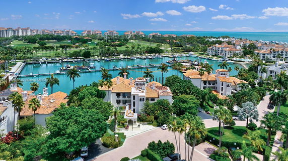 Aerial view of a waterfront residential area with buildings featuring terracotta roofs, surrounded by lush greenery and a marina.