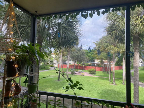View from a porch or balcony showing a grassy area with palm trees and a red fence, and a building in the background.