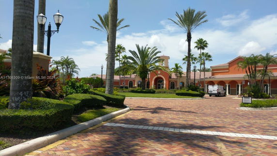 Front view of a large, single-story building with arched windows and a terracotta roof, surrounded by palm trees and landscaping.