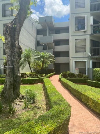 Front view of a multi-story residential building with balconies and a paved walkway leading through landscaped gardens.