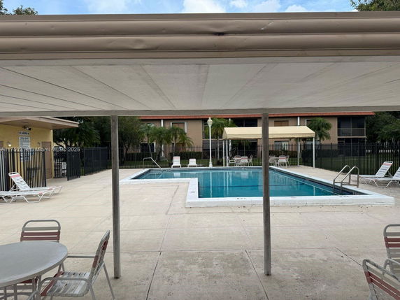 Outdoor swimming pool area with lounge chairs and tables, surrounded by a fence and nearby buildings.