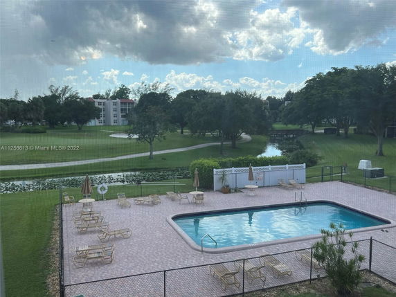 View of an outdoor swimming pool with lounging chairs in a fenced area, surrounded by greenery and buildings in the background.