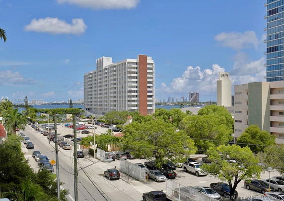 View of a multi-story building with balconies and a wide street lined with cars and trees, overlooking a coastal area.
