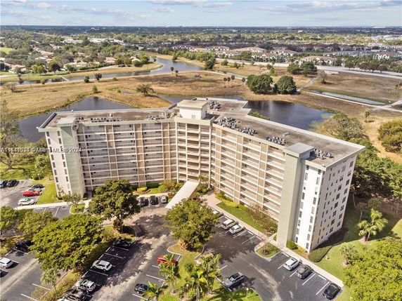 Aerial view of a multi-story residential building with parking area, surrounded by natural landscapes, water bodies, and distant residential areas.