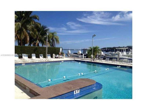 Outdoor swimming pool area overlooking a body of water with boats and palm trees.