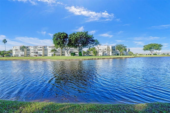Multiple four-story buildings with balconies, surrounded by trees, near a body of water.