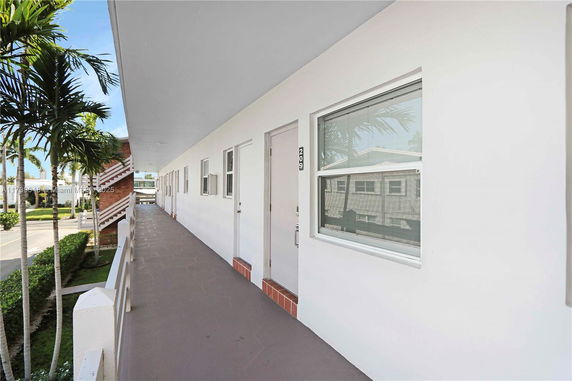 Exterior hallway of a building with white walls and multiple doors, surrounded by palm trees.