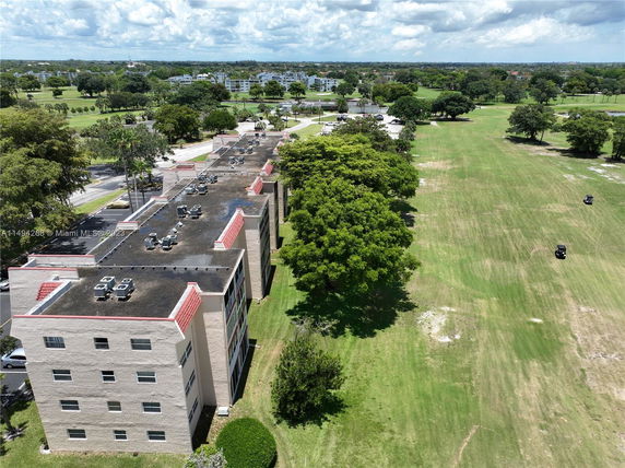 Aerial view of a row of apartment buildings with visible rooftops equipped with air conditioning units, set against a backdrop of open green spaces and trees.