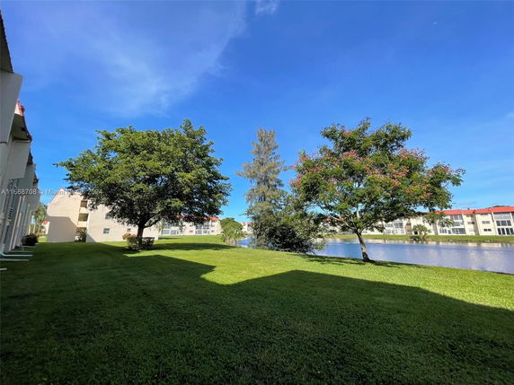 Wide view of grassy area with trees and a body of water, surrounded by buildings.