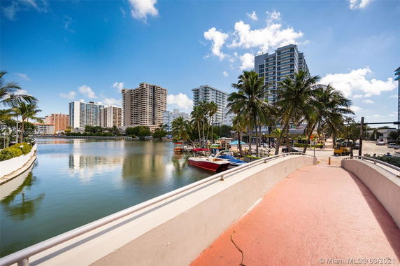 Scenic view of a waterfront area with high-rise buildings, palm trees, and moored boats under a clear blue sky.