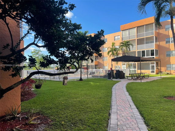 View of an apartment building with a grassy area and pathway leading to seating under a canopy.