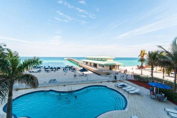 View of a beachside area with a swimming pool in the foreground and a pier extending into the ocean.