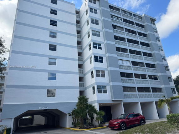 Front view of a multi-story residential building with balconies and a parking area at the entrance.