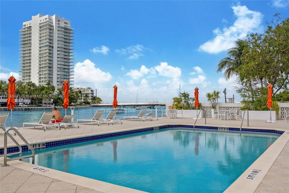 Outdoor swimming pool area with sun loungers and umbrellas, near a waterfront and a tall apartment building in the background.
