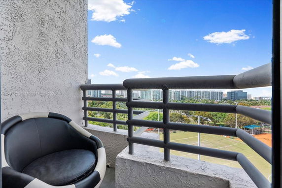 View from a high-rise balcony with metal railing and a chair, overlooking distant buildings and sports fields.