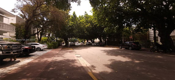 Outdoor parking area with several cars parked along the street under trees.
