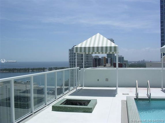Rooftop view with a small pool and a canopy structure. The area is surrounded by glass railings and overlooks distant buildings and a body of water.