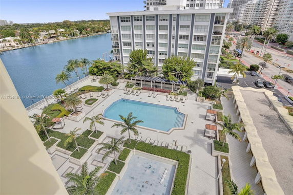 Aerial view of a swimming pool area with seating and landscaping, adjacent to a waterfront and high-rise building.
