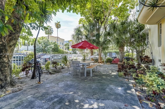 Outdoor seating area with table and chairs under a red umbrella, surrounded by trees and plants near a building.
