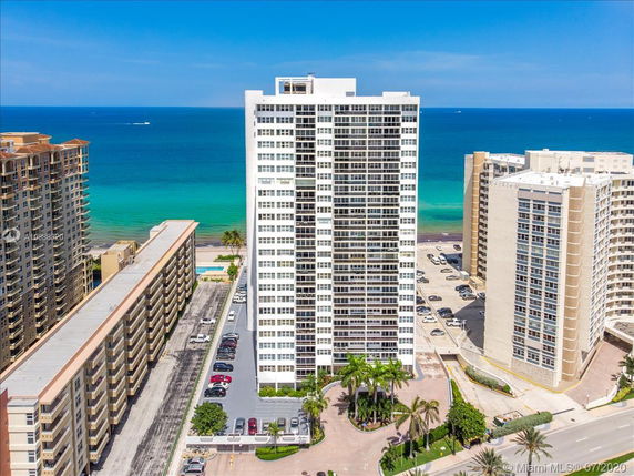 View of a tall residential building near the ocean with adjacent structures and parking areas.