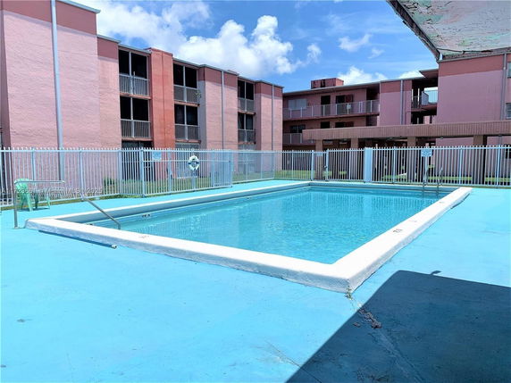 Outdoor swimming pool surrounded by a metal fence with a multi-story building in the background.