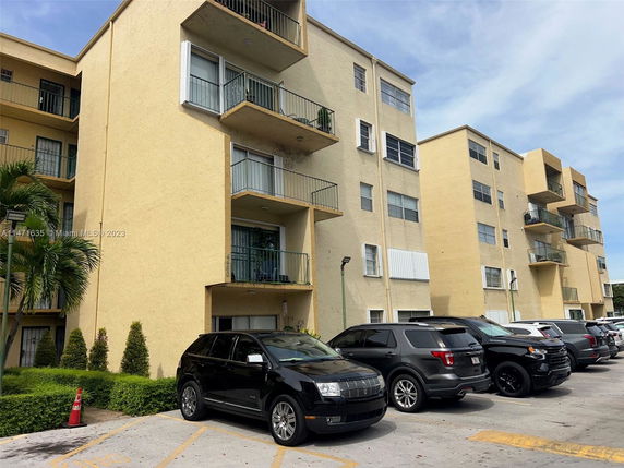 Front view of a multi-story residential building with balconies and adjacent parking area.