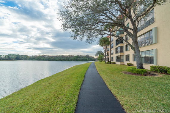View of a multi-story building with large windows next to a lake and a walking path.
