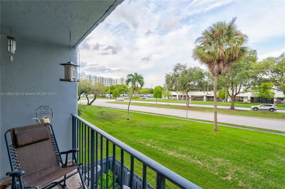 View from a balcony overlooking a street and green lawn with trees and buildings in the distance.