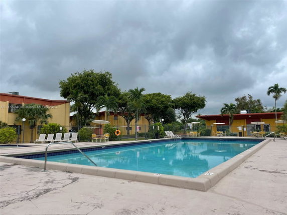 Swimming pool area with lounge chairs and umbrella-shaded tables, surrounded by trees and buildings.