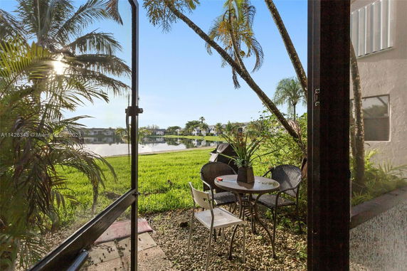 View from a patio showing a small outdoor seating area with a table and chairs, overlooking a lake and greenery.