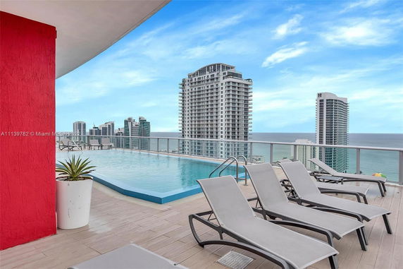 Rooftop with a pool and deck chairs overlooking the ocean and city skyscrapers.