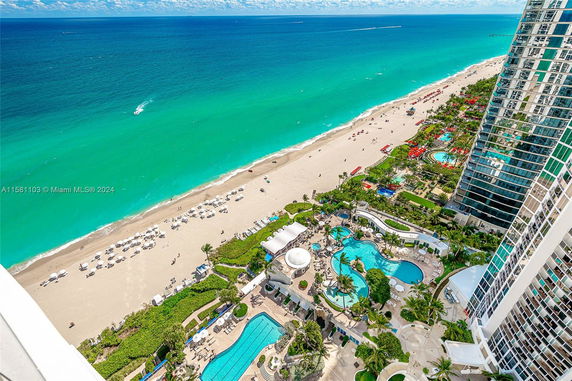 Panoramic view of a beachfront with clear blue water, sandy shores, and a large building with pools and landscaped areas.