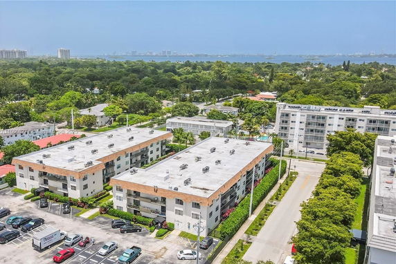 Aerial view of residential apartment buildings with a parking area and greenery, located near a body of water.