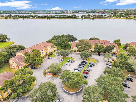 Aerial view of multiple residential buildings near a water body with a parking lot and surrounding greenery.