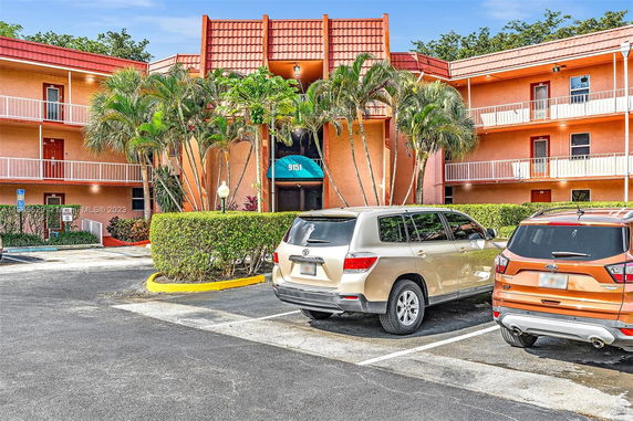 Parking area in front of a three-story building with balconies and tropical plants.