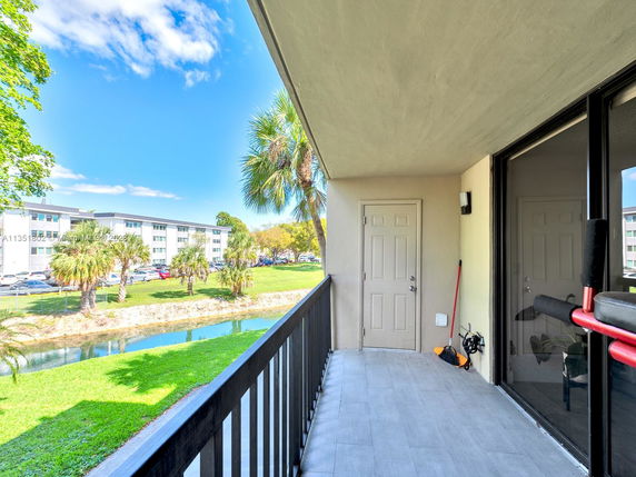 View from the balcony showing a grassy area, a small canal, palm trees, and a multi-story residential building.